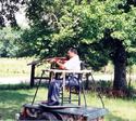 An adult male (with rifle aimed) is seated on his hunting stand (as described) that is atop the cargo bed of an old two-wheeled trailer.