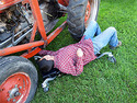 Laying on this creeper, an adult male is working on something under a tractor thatâ€™s parked on the grass.