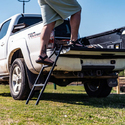 Back end of white pickup truck with tailgate down & black metal ladder attached to side of tailgate with man on ladder stepping onto tailgate