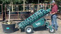 Man on right in blue jeans marron shirt and gray baseball cap guiding a 4-wheeled cart with the bed on about a 45 degree angle and a large potted tree is in front of the cart or transporter.